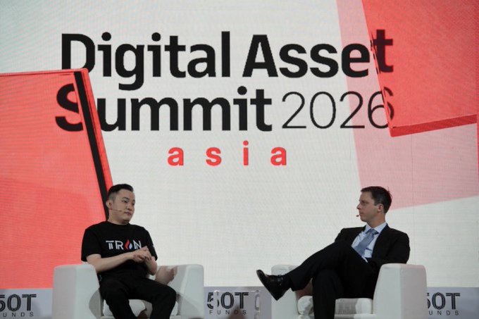 Two men sit on white chairs on stage at the 'Digital Asset Summit 2026 asia' conference; one wears a black 'TRON' shirt and the other wears a suit.