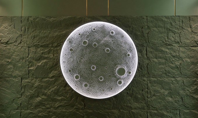 A high-resolution render of a cratered grey moon is centered against a background split between dark grey textured stone tiles on the bottom and smooth olive-green panels on top.