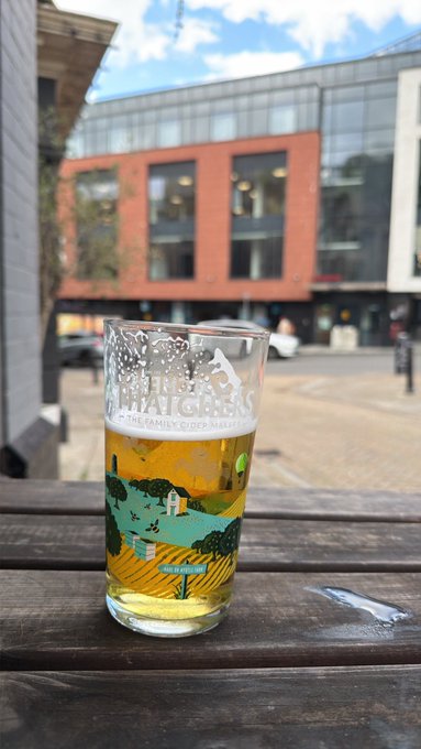 A pint glass filled with light beer sits on a wooden pub table, featuring a colorful landscape design and the text 'WHITGATE'S ALE THE FAMILY RUN BREWERY'.