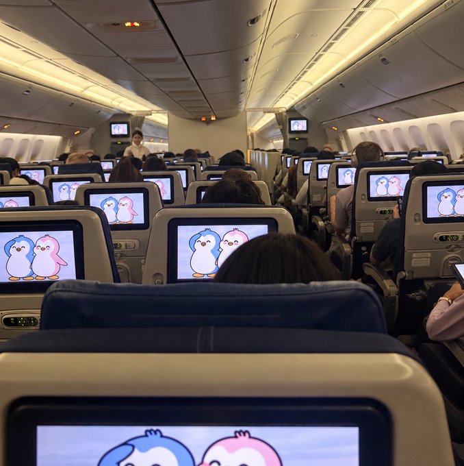 Interior of an airplane cabin showing passengers in seats with in-flight entertainment screens displaying two cartoon penguins.
