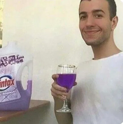 A man in a white shirt smiles while holding a wine glass filled with purple liquid, standing next to a purple bottle labeled 'Vintax'.