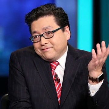 A man in a dark pinstripe suit with a red striped tie and glasses gestures with his hand raised while appearing on what looks like a television news set.