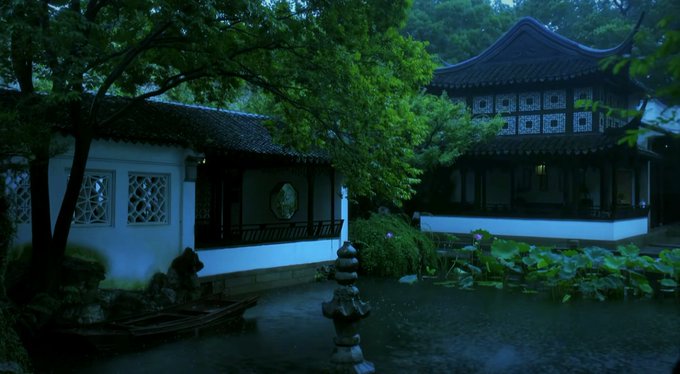 A serene, dimly lit traditional East Asian garden featuring a wooden pavilion, white-walled building with lattice windows, and a tranquil pond with lotus plants and a stone lantern in the foreground.