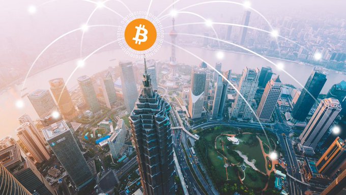 An aerial view of a city is overlaid with a Bitcoin logo and a glowing network of connected nodes, symbolizing blockchain activity. The graphic complements a crypto news roundup noting BTC near $63.6K, ETH around $1.81K, a $1.7M Maya Protocol exploit, Kalshi’s perpetual-futures plans, a $58M Singapore asset freeze, and a prediction that BTC could return to $100K.