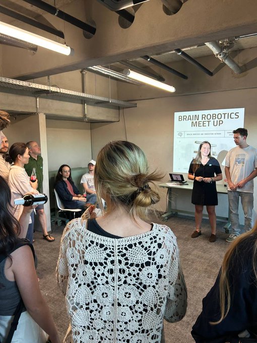 A photo taken at a 'Brain Robotics Meet Up' event showing a woman in a dark dress presenting to a crowd in an industrial-style room, with a slide visible in the background.