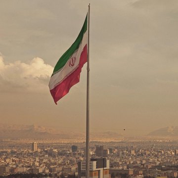 The Iranian flag flies over a sprawling city skyline with mountains in the distance. The image relates to the post by visually representing Iran, the country whose central bank, IRGC, and gambling sites were linked to the Binance-bound crypto exchange Shelbit.