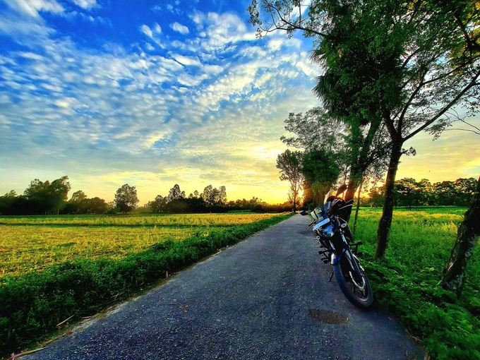 A scenic sunset photo showing a motorcycle parked on a narrow paved path between lush green rice fields and trees under a dramatic blue sky with scattered clouds.