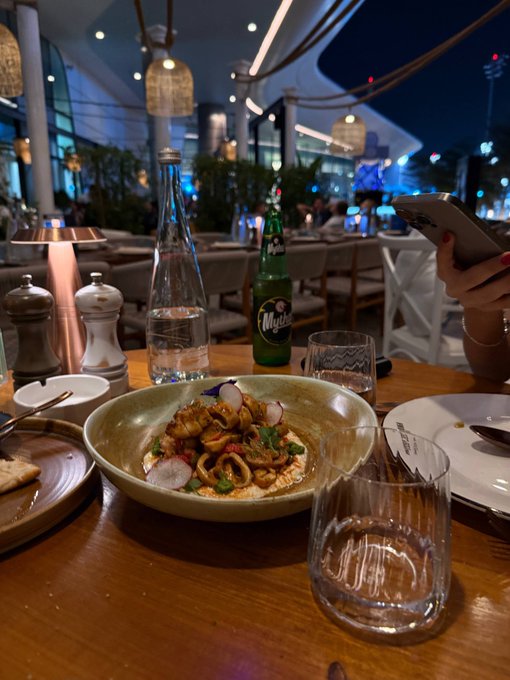 A restaurant table scene featuring a bowl of pasta or seafood dish, a bottle of Perrier water, a green sparkling beverage, salt and pepper shakers, and a person's hands holding a phone.
