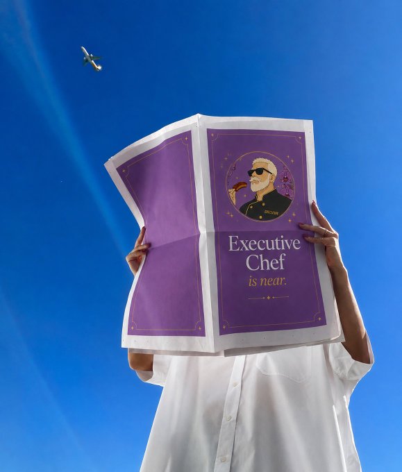 A person in a white shirt holds up a large purple menu or booklet reading "Executive Chef is near" featuring an illustration of a chef, photographed against a blue sky with an airplane overhead.
