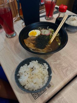 A wooden restaurant table holds a bowl of ramen with an egg and nori, a side bowl of rice, chopsticks, and glasses of a red beverage.
