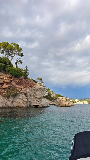 A scenic view from a boat on turquoise water showing rocky cliffs covered with green trees, with white buildings nestled into the hillside under a cloudy sky.