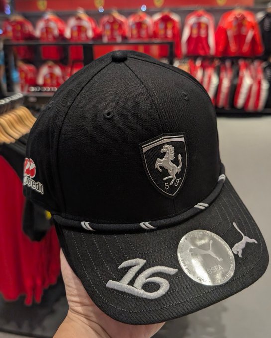 A black Ferrari Scuderia cap with the number 16, featuring a Puma logo, held in front of a display of red Ferrari apparel.