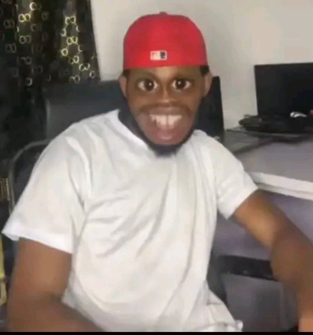 A man in a red MLB cap and white t-shirt sits at a desk, making an exaggerated open-mouthed expression at the camera.