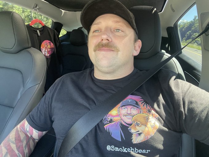 A man wearing a black cap and a "@Smokeabear" t-shirt featuring a cartoon portrait of himself and a dog sits in the driver's seat of a Tesla with a black t-shirt hanging in the backseat. The image captures him tagged to Nicki Minaj ahead of "The Pinkprint 10/10" takeover event, likely teasing a merch or collaboration drop.