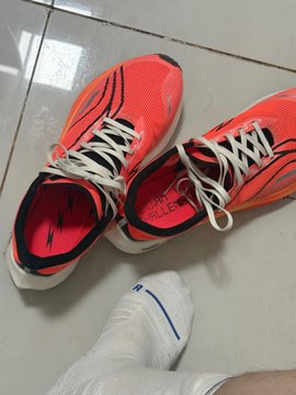 A pair of bright orange and white athletic running shoes (appearing to be ASICS brand based on the visible logo) sits on a tiled floor next to a white sneaker, photographed from above with someone wearing white socks nearby. The image's vibrant, energetic color palette and fresh appearance visually contrast with the post's darkly humorous "Dead dog, crawl over here" caption, creating a jarring juxtaposition between the lively shoes and the morbid phrase.