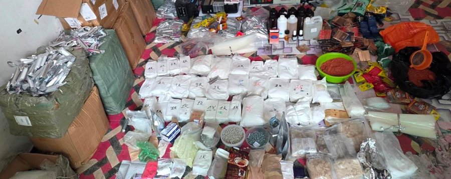 A large assortment of suspected drug-manufacturing chemicals, powders, pills, and equipment is spread across a floor, including bagged substances and assorted bottles. The seized materials relate to a DEA operation with Cambodian authorities targeting Mexican and Pakistani networks allegedly manufacturing methamphetamine and fentanyl and laundering money through cryptocurrency, with links to the Sinaloa Cartel.
