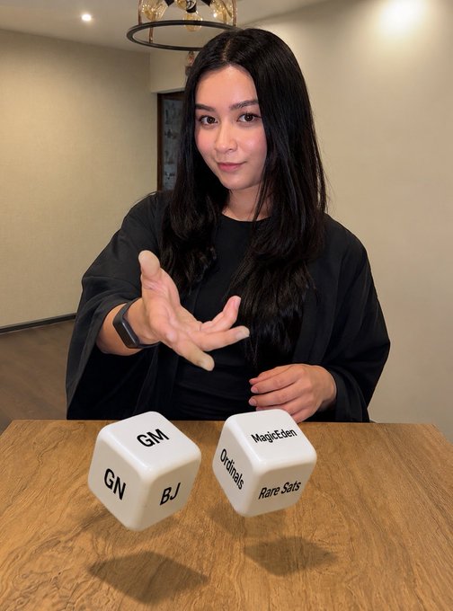 A woman in a black outfit reaches out to roll two large white dice on a wooden table, with the dice displaying abbreviations like 'GM', 'GN', 'BJ', 'Ordinals', 'MagicEden', and 'Rare Sats'.