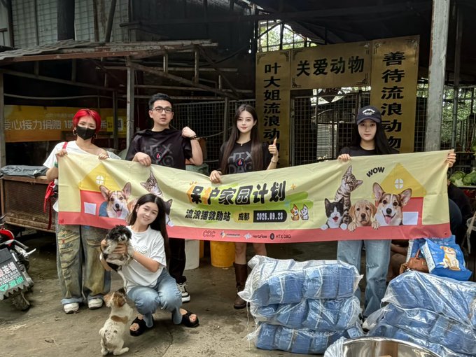 A group of five people poses with a banner reading 'LEO P家园计划' at an animal shelter, surrounded by rescued dogs and stacks of blue pet food bags in front of kennels.