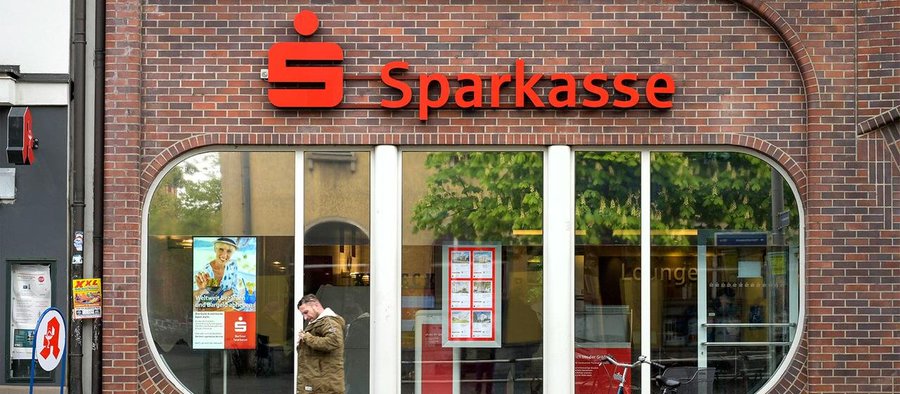 The image shows the storefront of a Sparkasse bank branch, featuring its distinctive red "S" logo and signage mounted on a brick facade above arched windows. The accompanying text highlights how Sparkassen, Germany's vast banking network, is now integrating cryptocurrencies like BTC, ETH, XRP, SOL, and POL into its app, representing a significant step in bringing digital assets into mainstream traditional banking.