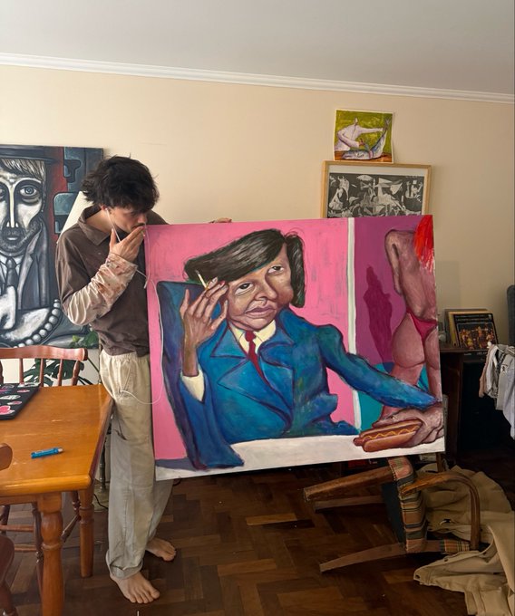 A photograph of a barefoot man in a brown shirt standing next to a large, vibrant painting of a man in a blue suit smoking a cigarette, with a partial nude figure visible on the canvas.