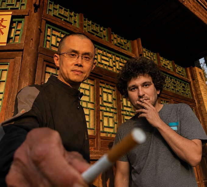A photo of two men; one in a black athletic top extends a cigarette toward the camera, standing beside a curly-haired man in a grey t-shirt who is smoking, in front of an ornate wooden window.