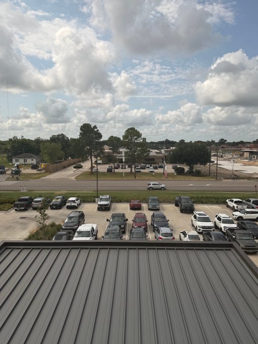 A view from the elevated vantage of the cancer center overlooks a busy parking lot filled with cars, a passing minivan on the road, and surrounding suburban buildings under a partly cloudy sky. The metal roof in the foreground suggests the photo was taken from an upper floor of the medical facility.