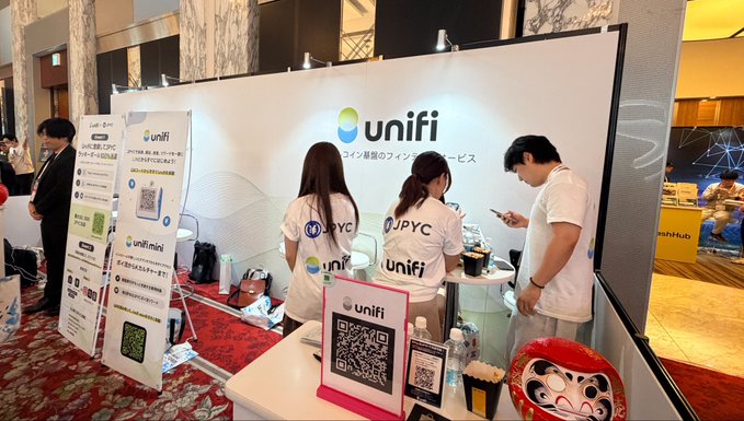 A photo taken at the Unifi booth, showing staff in branded "unifi" and "JPYC" shirts engaging with a visitor at the counter, alongside promotional materials featuring the Unifi logo and QR codes. The image reflects the post's enthusiasm for both the Unifi service and a personal investment in JPYC, highlighting the brand's active presence at the event.