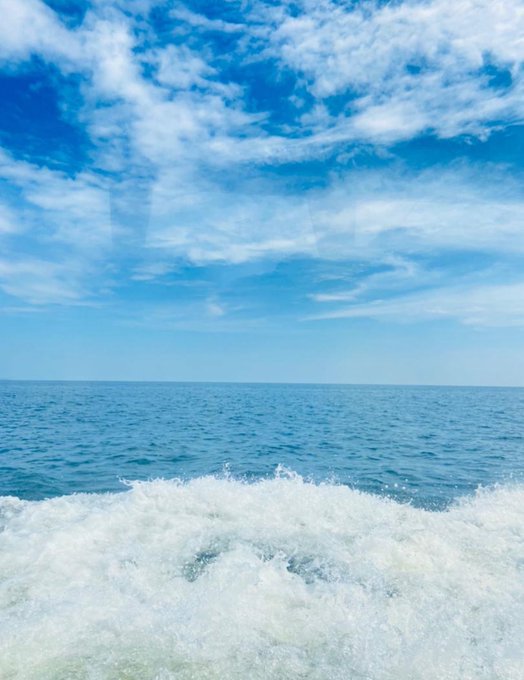 A view from the back of a boat shows churning white wake against a vast blue ocean and partly cloudy sky.