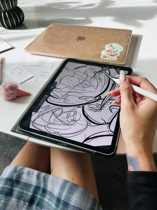 A person's hands using a white stylus to draw on a tablet, with a laptop, ice cream cone decoration, and notes visible on a white desk.