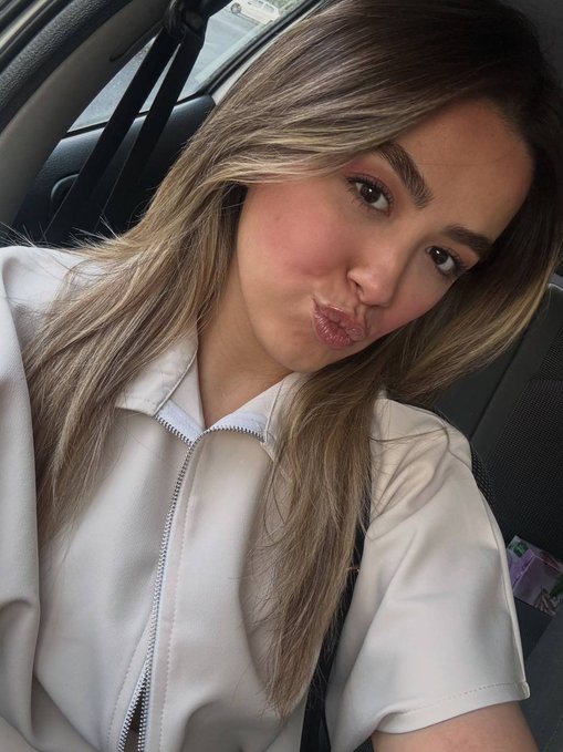 A selfie of a young woman with long highlighted hair making a kissing face, wearing a cream-colored zip-up jacket while sitting inside a car.