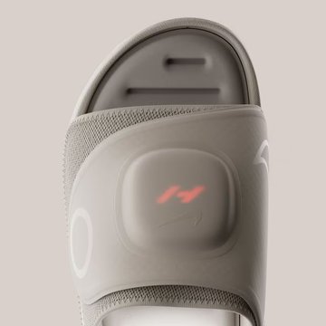 A close-up product shot of a gray Nike slide sandal with an illuminated red 'M' logo on the adjustable strap.