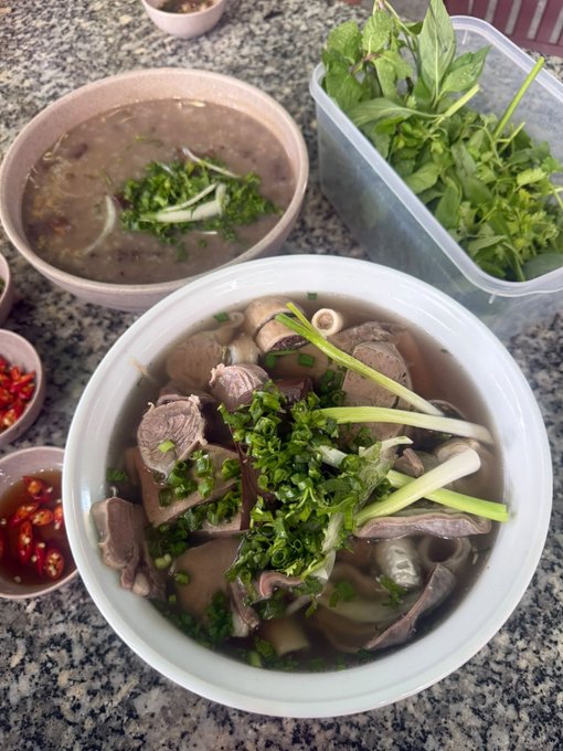 A steaming bowl of Vietnamese phoêu, garnished with fresh herbs, chopped scallions, and accompanied by chili sauce, sits alongside a smaller bowl of broth. The image doesn't directly relate to the morning greeting and crypto project mentions in the accompanying post, instead showcasing a hearty meal, possibly to thank supporters or illustrate a daily routine.