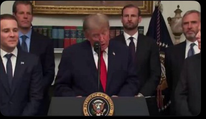 Donald Trump speaking at a podium with the presidential seal, surrounded by several men in suits in what appears to be the Oval Office.