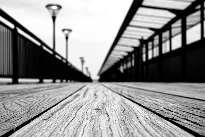 A black and white low-angle photograph looking down a long wooden boardwalk, with railings and lampposts stretching into the distance under a cloudy sky.