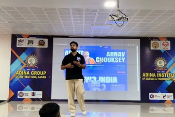 A man stands on stage at Adina Institute of Science, Technology & Management, giving a presentation with a slide introducing 'ARNAV CHOUKSEY' displayed behind him.