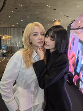 Two young women pose closely together indoors; one with long blonde hair in a white shirt and another with dark hair in a black jacket, with the blonde touching the other's chin, set against a blurred indoor backdrop with a chandelier and a colorful display screen.