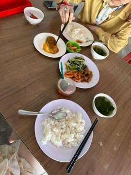 An overhead view of a shared Chinese meal at a wooden table with rice, vegetables, and dumplings, with a person using chopsticks.