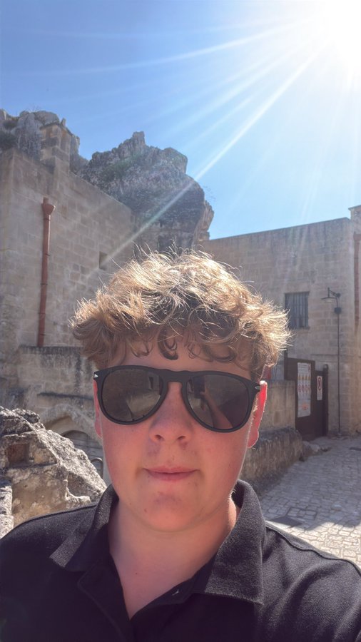 A young man with curly hair and sunglasses takes a selfie in front of ancient stone ruins under a bright sunny sky, fitting the confident "Quant kid is back" vibe of the post as he positions himself as a leader of "Solana summer." No visible text, tickers, or branding appear in the image to reinforce the $QUANT promotion.