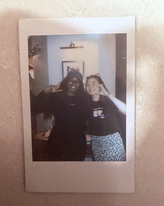 A Polaroid photo shows two smiling women posing together indoors, one in a black outfit and cap, the other in a black tee and leopard-print skirt, both making peace signs. The casual, friendly snap captures the wholesome, close-knit vibe highlighted in the post about Superteam Thailand's welcoming community.