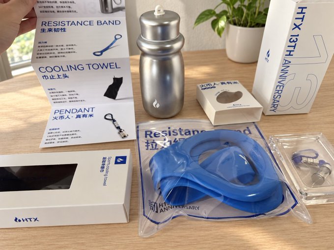 A silver Huobi HTX 13th Anniversary merchandise set is displayed on a wooden desk, featuring a stainless steel water bottle, a blue resistance band labeled "Resistance Band 拉力器," a cooling towel, and a pendant keychain, alongside packaging marked "HTX 13TH ANNIVERSARY" and a "Smart Digital Display" box.