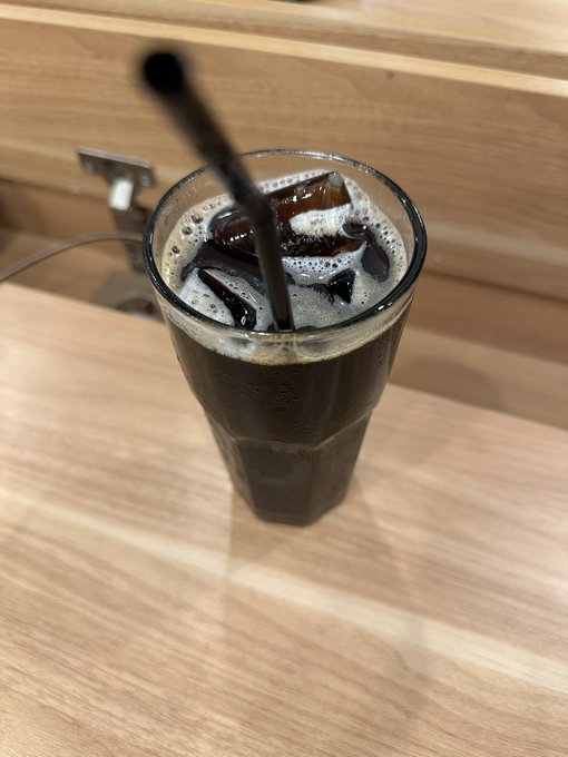 A tall glass of dark iced coffee with foam sits on a light wooden counter, with a black straw rising from the drink.