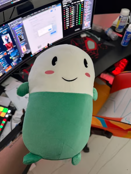 A plush toy of $CUPSEY, the popular meme coin mascot, is held up in front of a trading setup with multiple monitors displaying market data and charts. The image embodies the post's sentiment, showcasing the iconic smiling green-and-white character as the unofficial welcome figure for new traders entering the crypto markets.