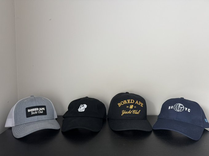 Four baseball caps lined up on a dark surface, including three black "BORED APE Yacht Club" caps in different styles and one navy cap with a globe logo.