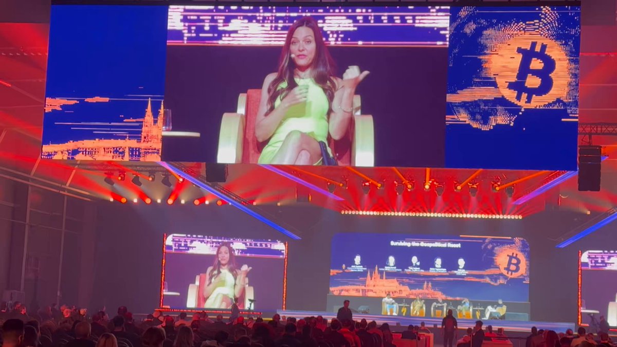 The image shows a stage with a large screen displaying an event or presentation related to Bitcoin. The screen features a woman sitting in a chair, holding a cat, which appears to be the main focus of the presentation. There are also other people present on the stage and around it, suggesting that this is a public event where various individuals have gathered to learn about Bitcoin and its implications.