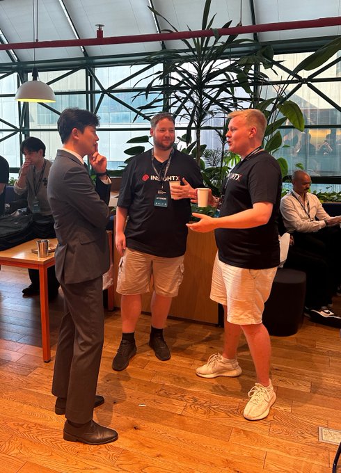 Three men converse in a modern, glass-roofed room; one wears a suit while the other two wear black 'INSIGHTS' t-shirts. Plants and other people are visible in the background.