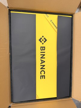 The image features an open cardboard box containing a black and yellow item with the word "Binance" written on it. The box appears to be from Binance, as indicated by the text on the item inside. The box is placed on top of a brown surface, possibly a table or countertop.