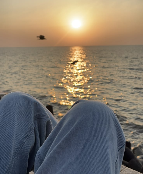 A first-person view shows someone wearing blue jeans sitting by the ocean at sunset, with the sun reflecting on the water and a bird flying in the distance.