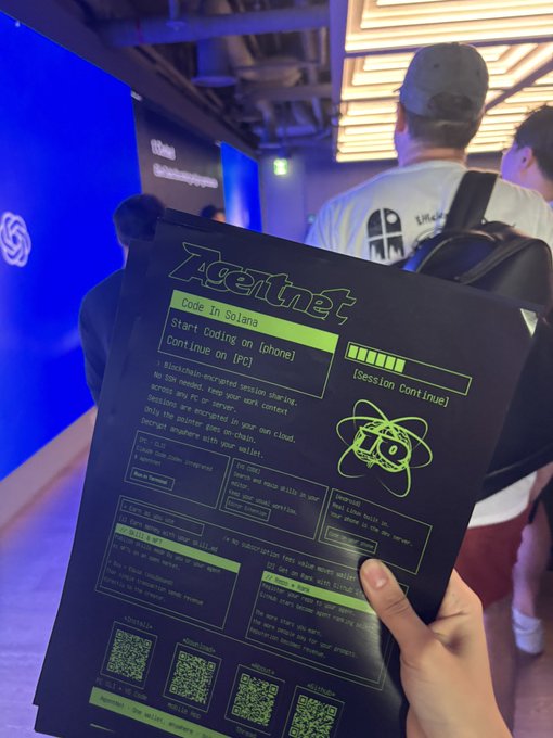 A hand holds up an "Igetnet; Code in Solana" flyer at a tech meetup, which matches the post about shilling AgentNet at the Codex event. The flyer details instructions for coding on PC or phone, sessions continued "1D," and lists multiple QR codes for session details and the AgentNet website.