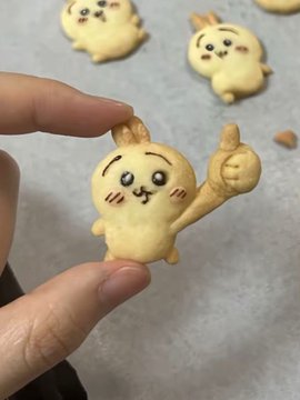 A hand holds up a small, cute baked pastry shaped like a cartoon character with a smiling face and thumbs up gesture, with similar baked figures visible on the parchment paper behind it.