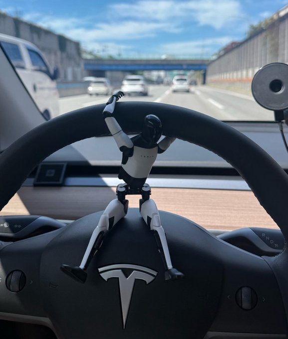 A small humanoid robot figurine is positioned on the steering wheel of a Tesla vehicle while driving on a highway under a blue sky.
